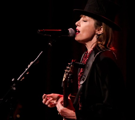 Une femme de profil, portant du noir et un chapeau haut de forme, tenant une guitare, chante dans un micro.